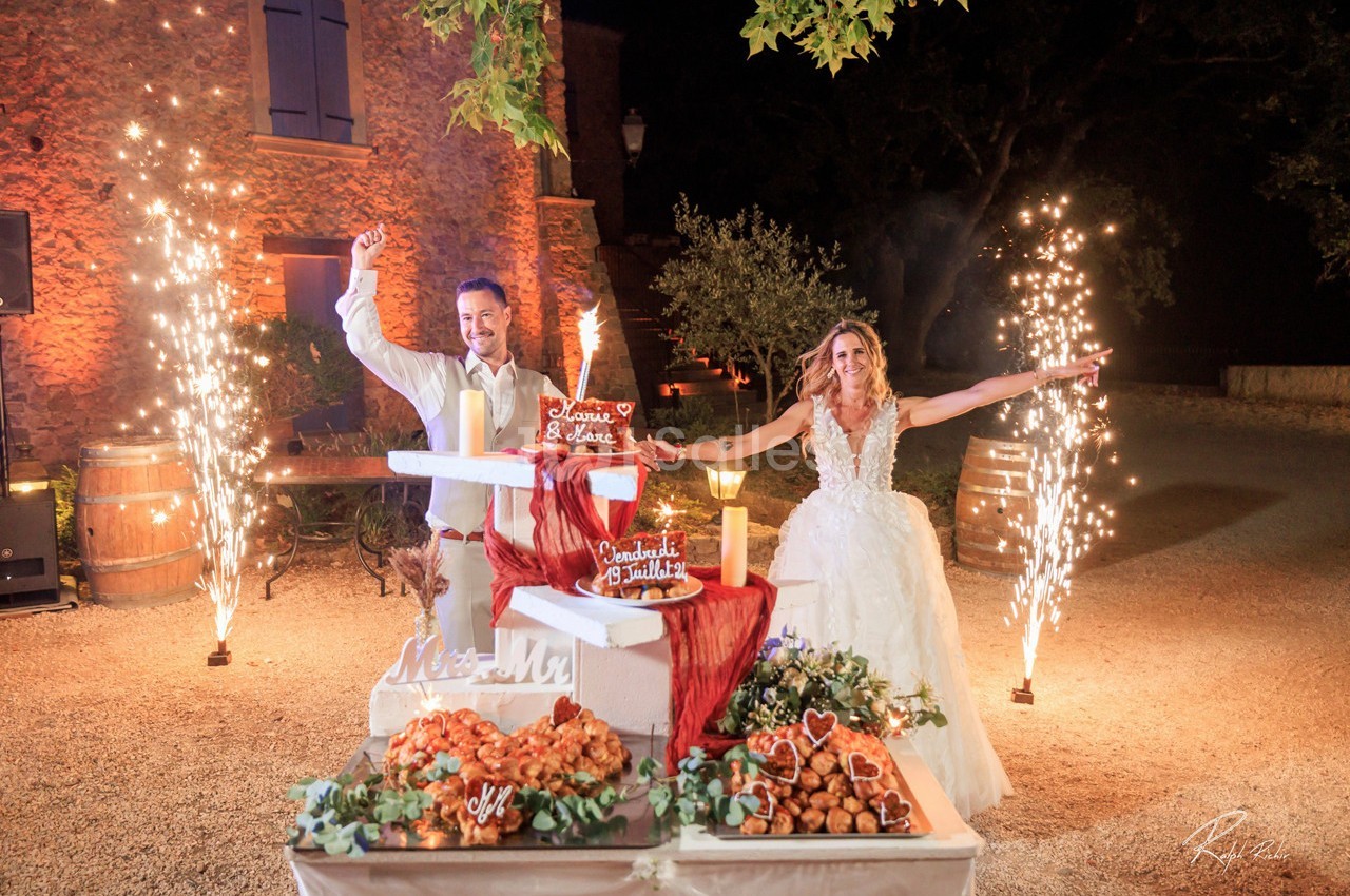 Un couple en tenue de mariage célèbre devant une pièce montée illuminée par des feux d'artifice.
