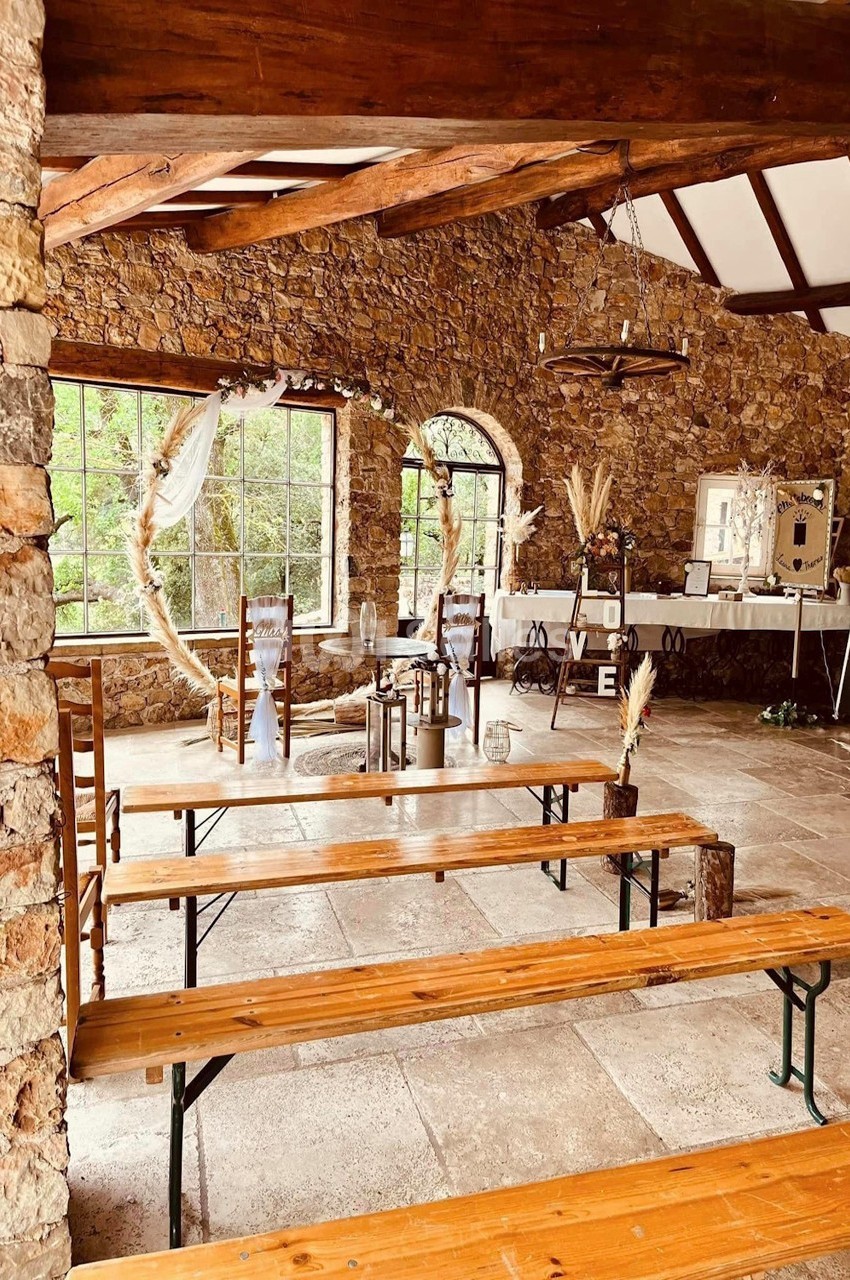 Salle en pierre avec bancs en bois, décor champêtre, grandes fenêtres et table ornée pour un événement.