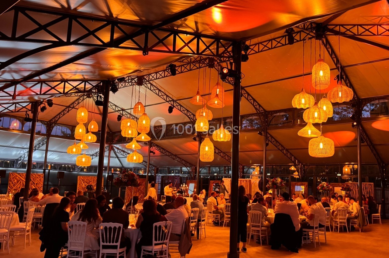 Salle de réception illuminée par des suspensions lumineuses, avec des invités assis à des tables rondes.
