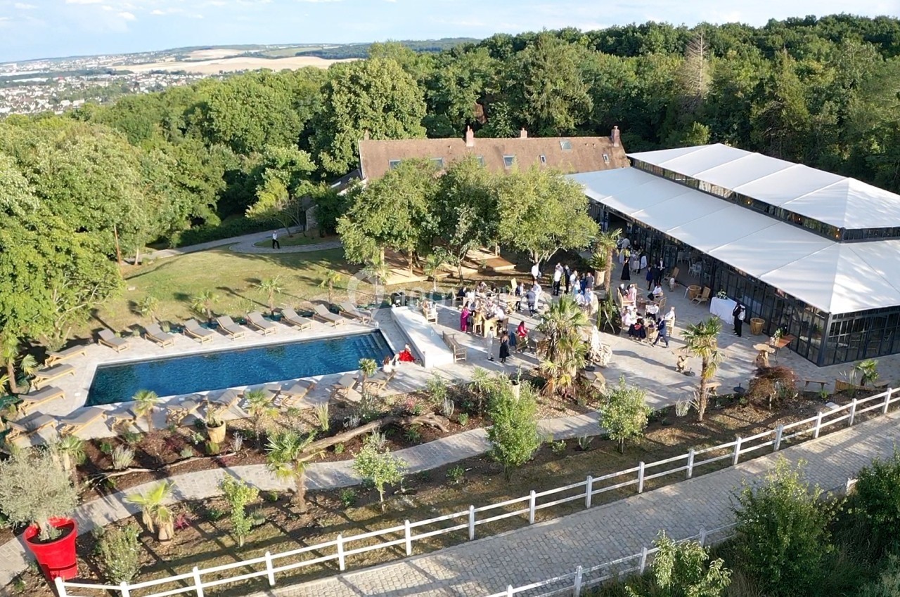 Vue aérienne d'un domaine avec piscine, terrasse animée par des invités, jardin arboré et bâtiment principal en arrière-plan.