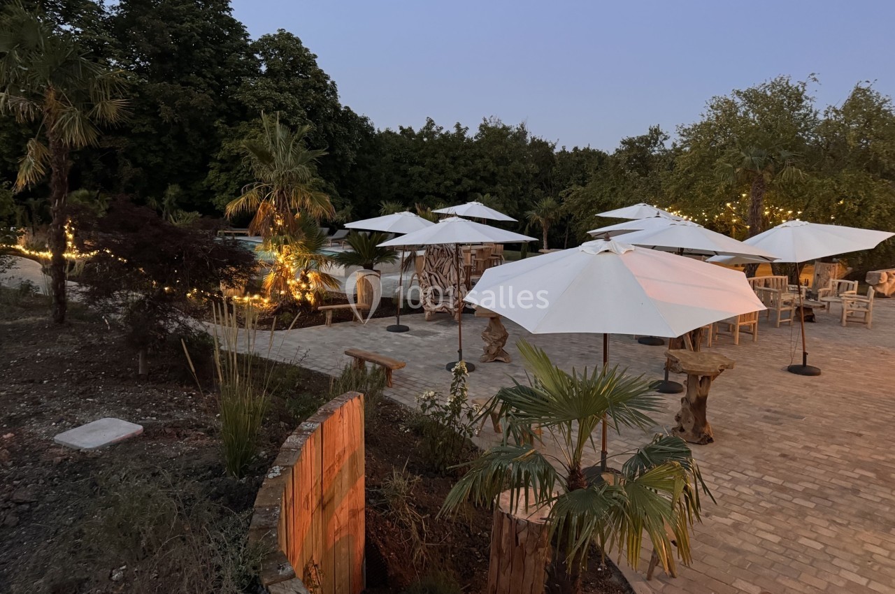 Terrasse pavée avec tables, chaises et parasols blancs entourée de végétation et éclairée par des guirlandes lumineuses.