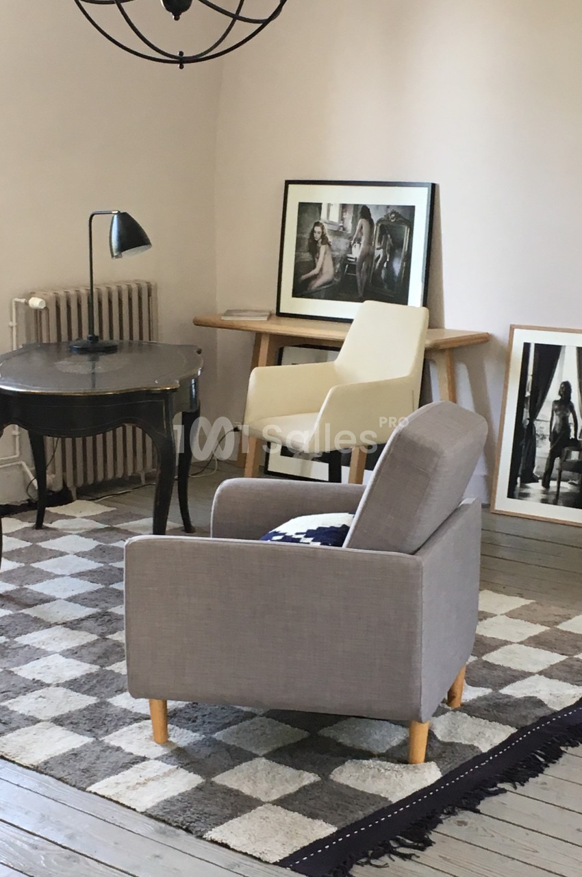 Salon avec fauteuils, bureau noir, lampe de table et cadres posés sur un meuble en bois, tapis à motifs au sol.