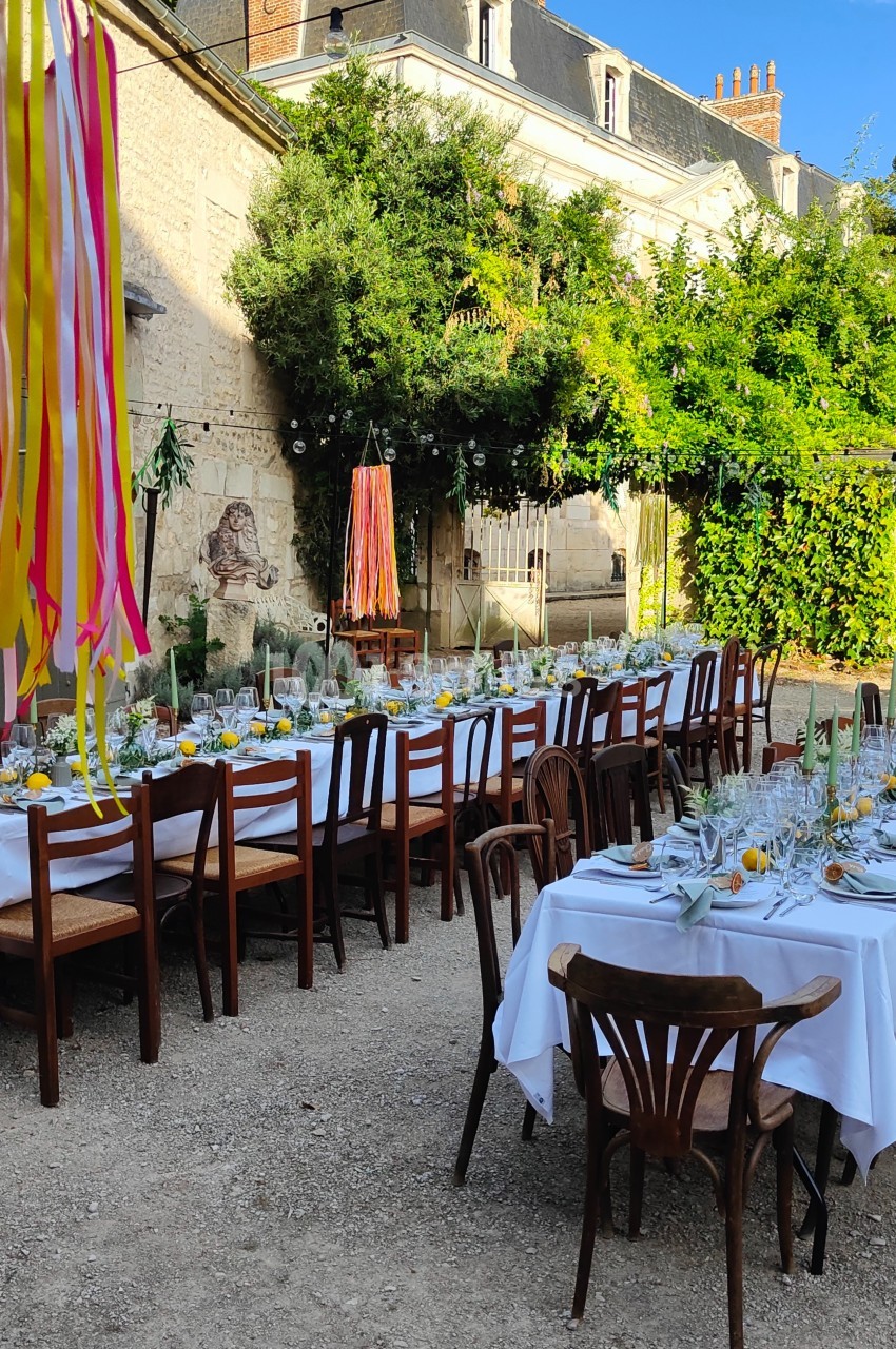 Tables dressées avec nappes blanches, décorées de fleurs et citrons, dans une cour extérieure bordée de verdure.