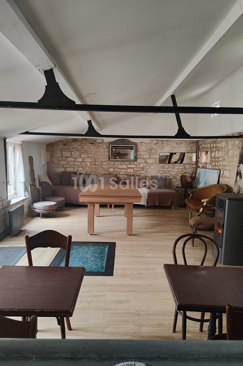 Salle lumineuse avec murs en pierre, tables en bois, poêle à bois et canapé au fond sous des poutres apparentes.