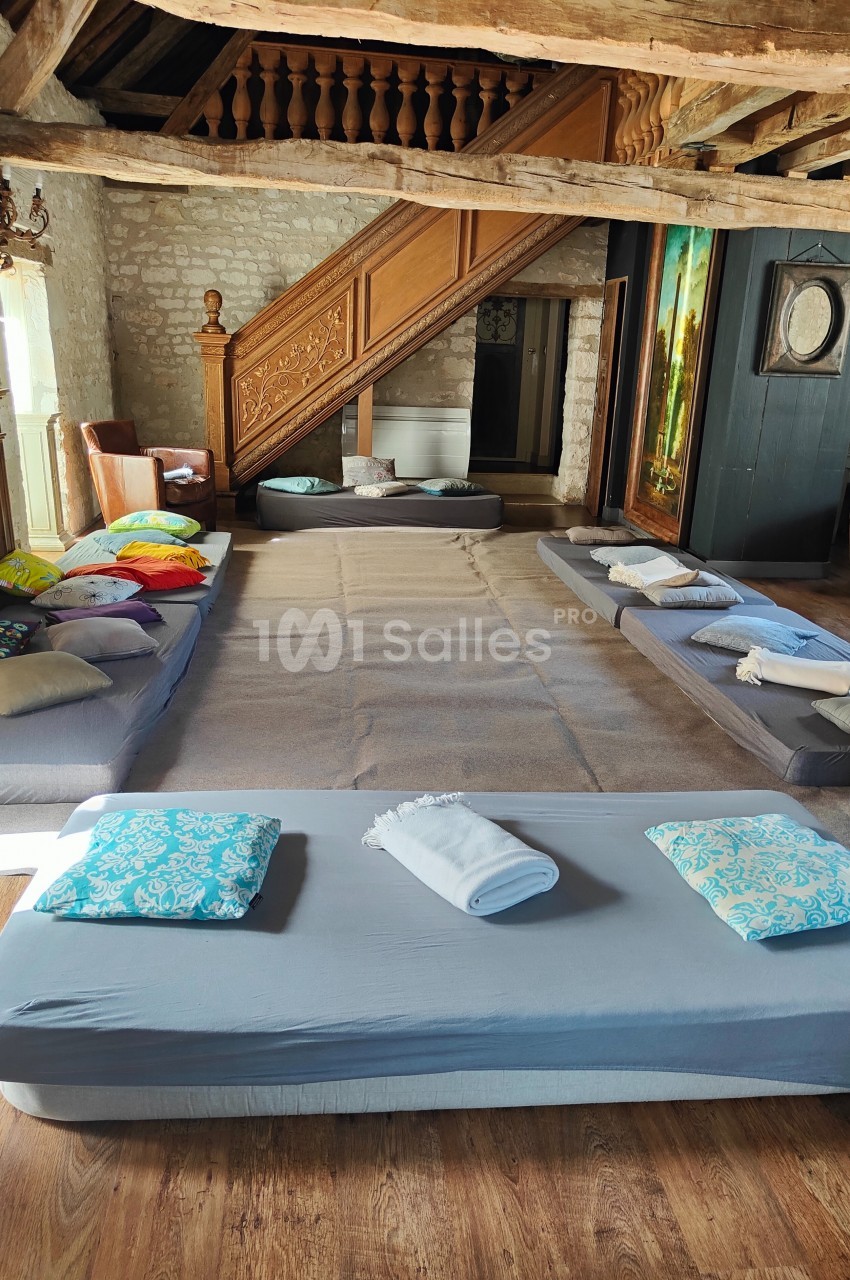 Matelas et coussins disposés au sol dans une pièce rustique avec poutres apparentes et escalier en bois sculpté.