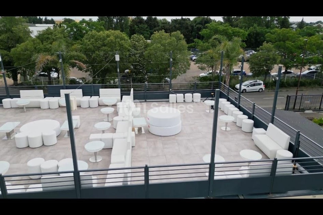 Terrasse aménagée avec mobilier blanc moderne, entourée de verdure et donnant sur un parking en arrière-plan.