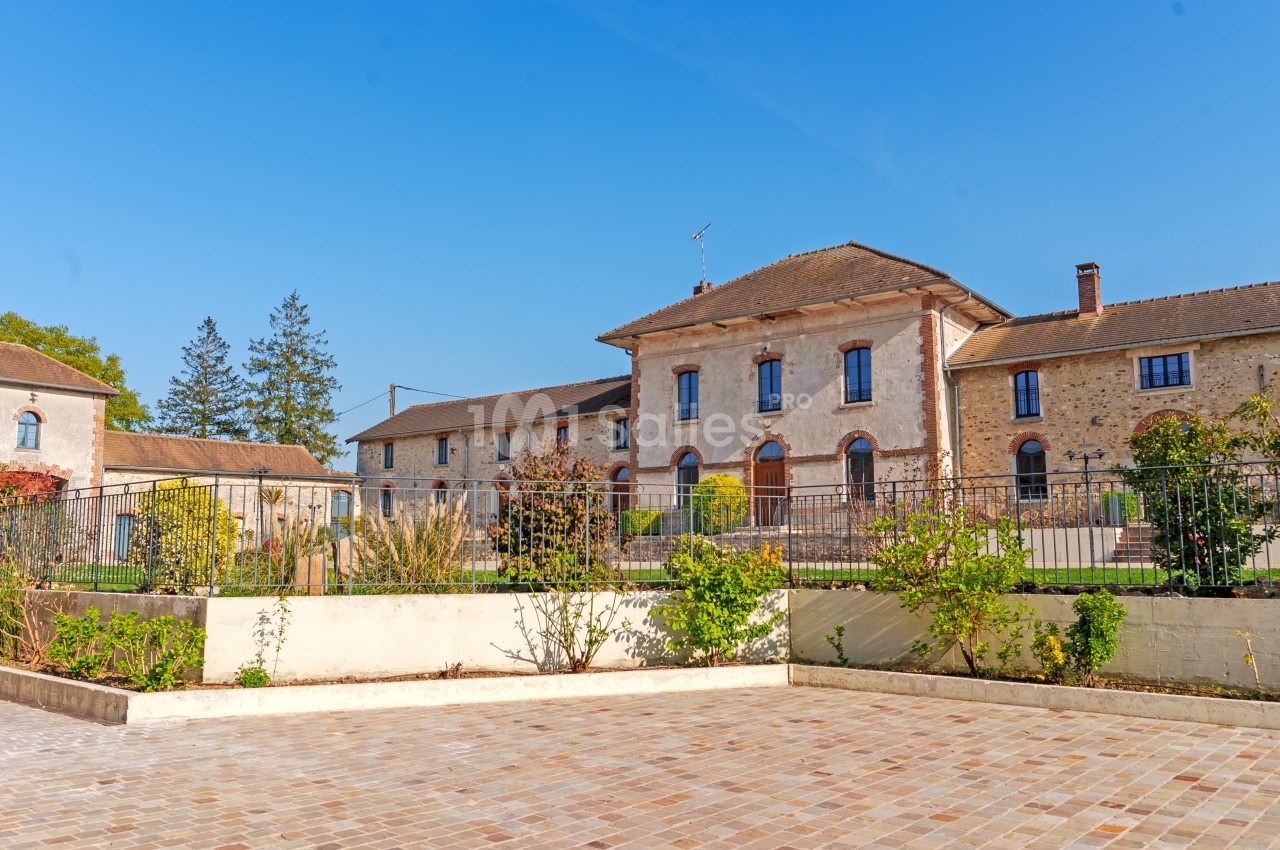 Bâtiment ancien en pierre avec toit en tuiles, entouré d'un jardin et d'une cour pavée sous un ciel bleu.