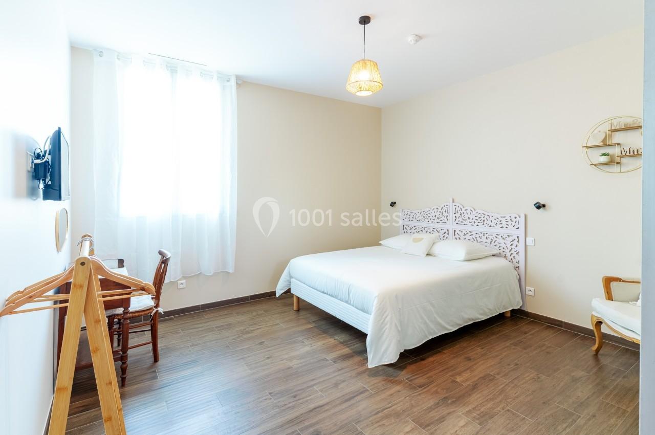 Chambre lumineuse avec lit double, parquet, mur beige, chaise, portant en bois et fenêtre avec rideaux blancs.