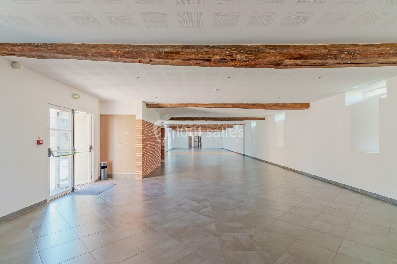 Salle vide avec sol en carrelage, poutres apparentes au plafond, murs blancs et lumière naturelle entrant par les fenêtres.