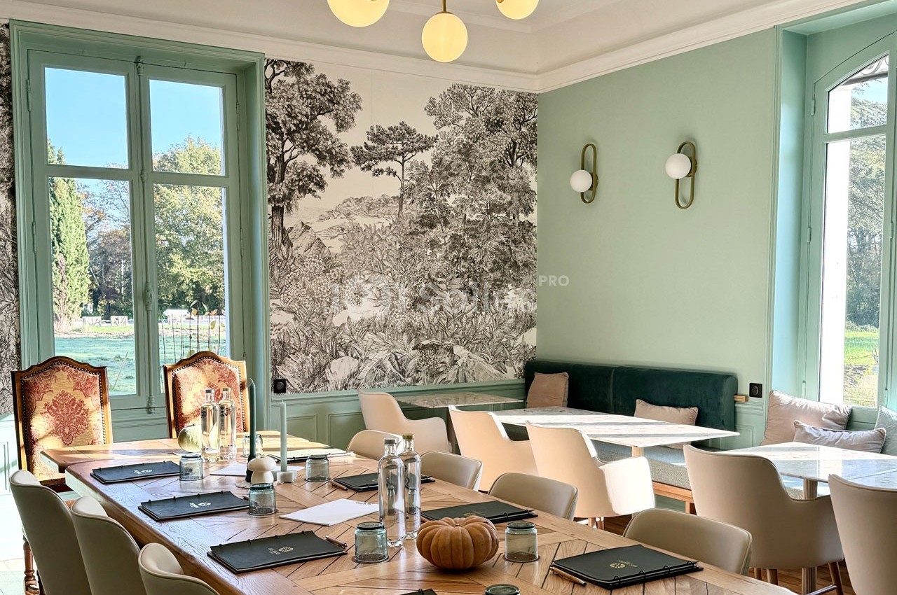 Salle lumineuse avec une grande table en bois, chaises modernes, fresque murale et vue sur un jardin à travers les fenêtres.