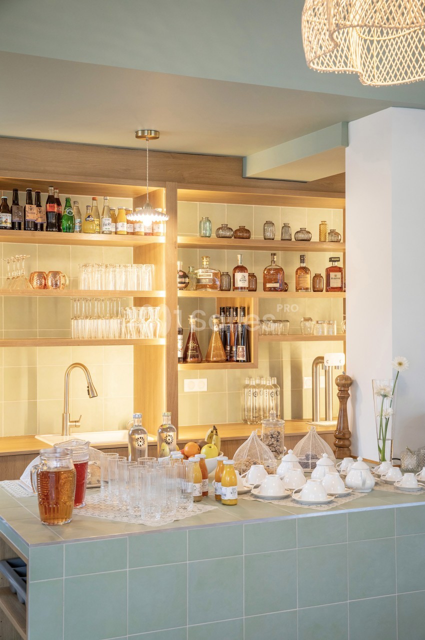 Un comptoir de cuisine avec boissons, verres, théières et étagères remplies de bouteilles en arrière-plan.