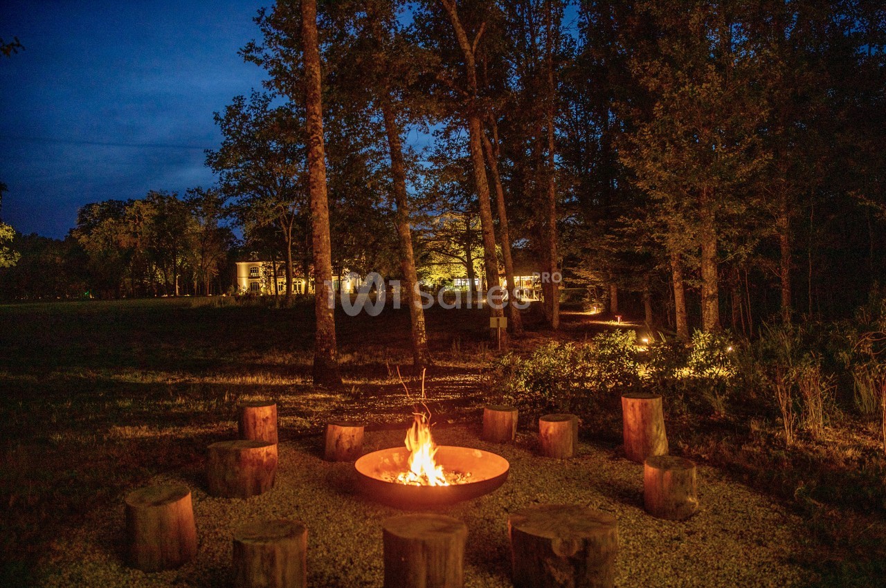 Feu de camp entouré de rondins de bois dans une clairière boisée, avec des lumières de maison en arrière-plan.
