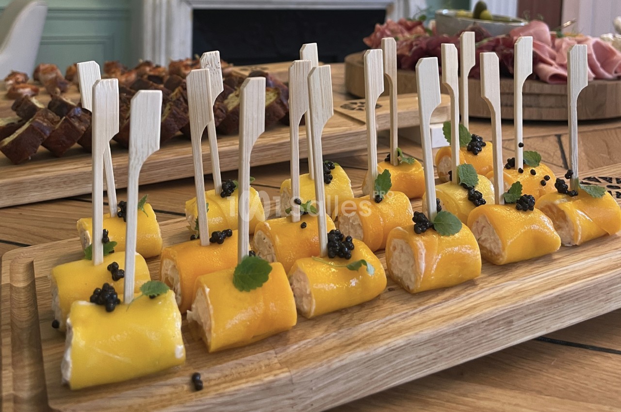 Plateau de bouchées roulées au fromage et mangue, décorées de caviar et de feuilles, sur une table en bois.
