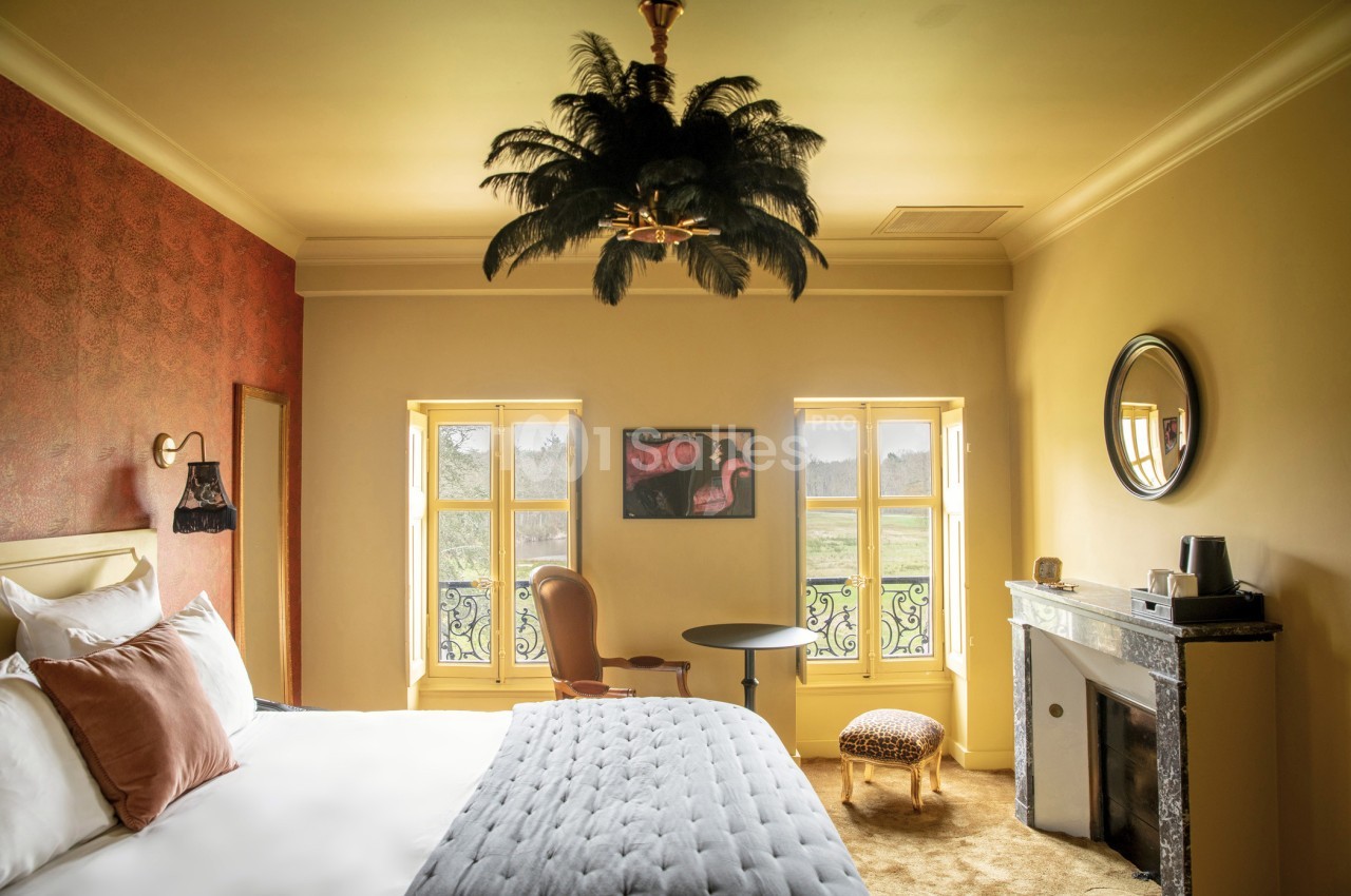 Chambre lumineuse avec lit double, fauteuil, table, cheminée, et vue sur un paysage extérieur à travers des fenêtres.