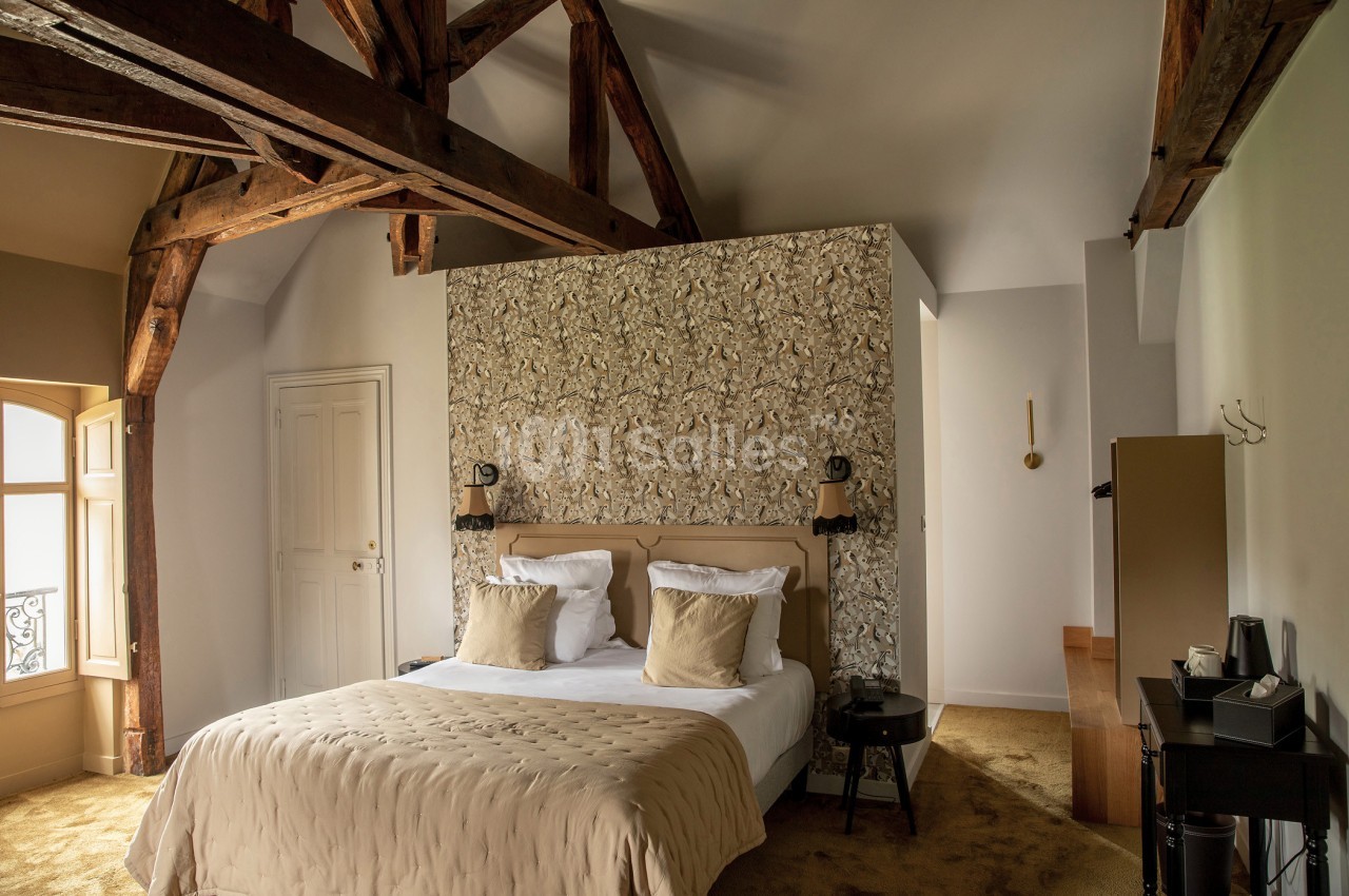 Chambre lumineuse avec poutres apparentes, lit double, tête de lit décorative et mobilier moderne.