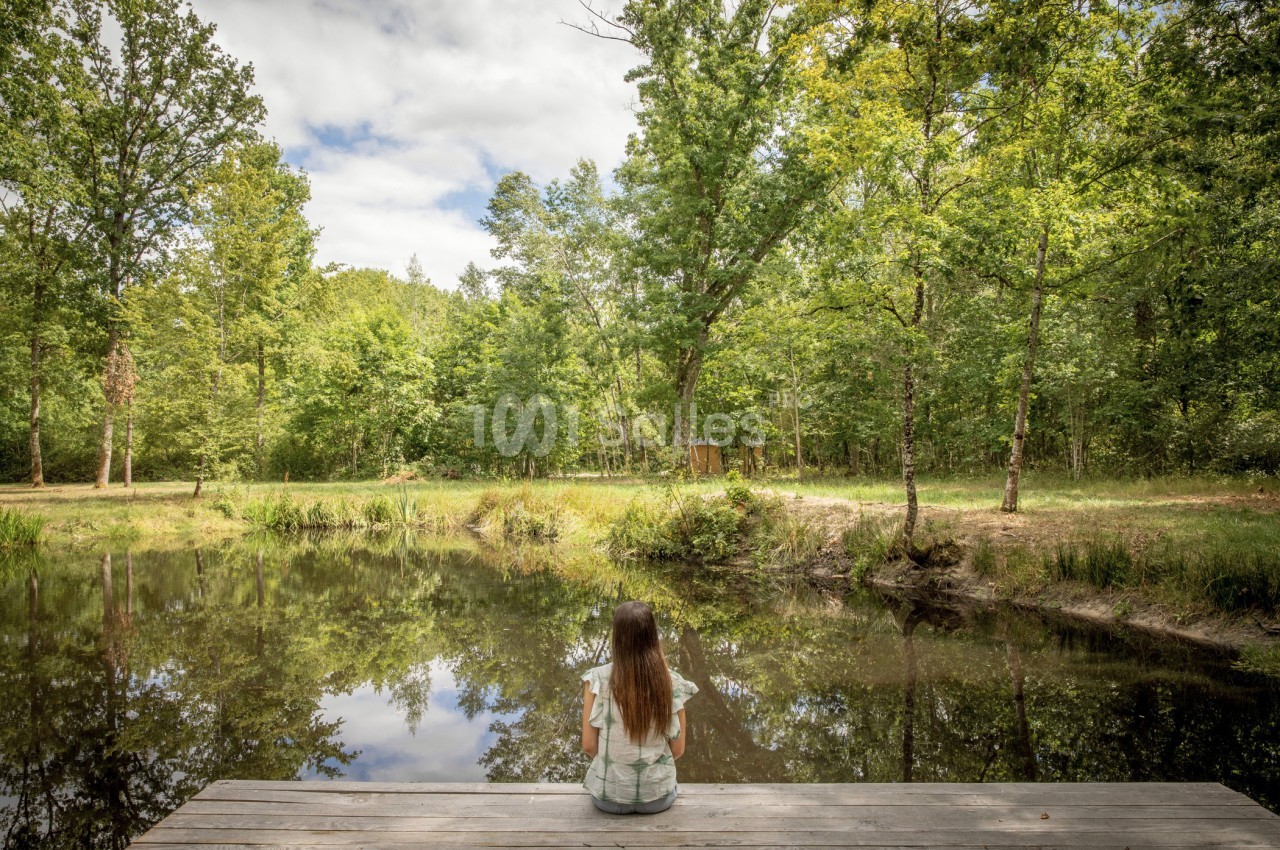Une femme assise sur un ponton en bois contemple un étang entouré d'arbres sous un ciel partiellement nuageux.