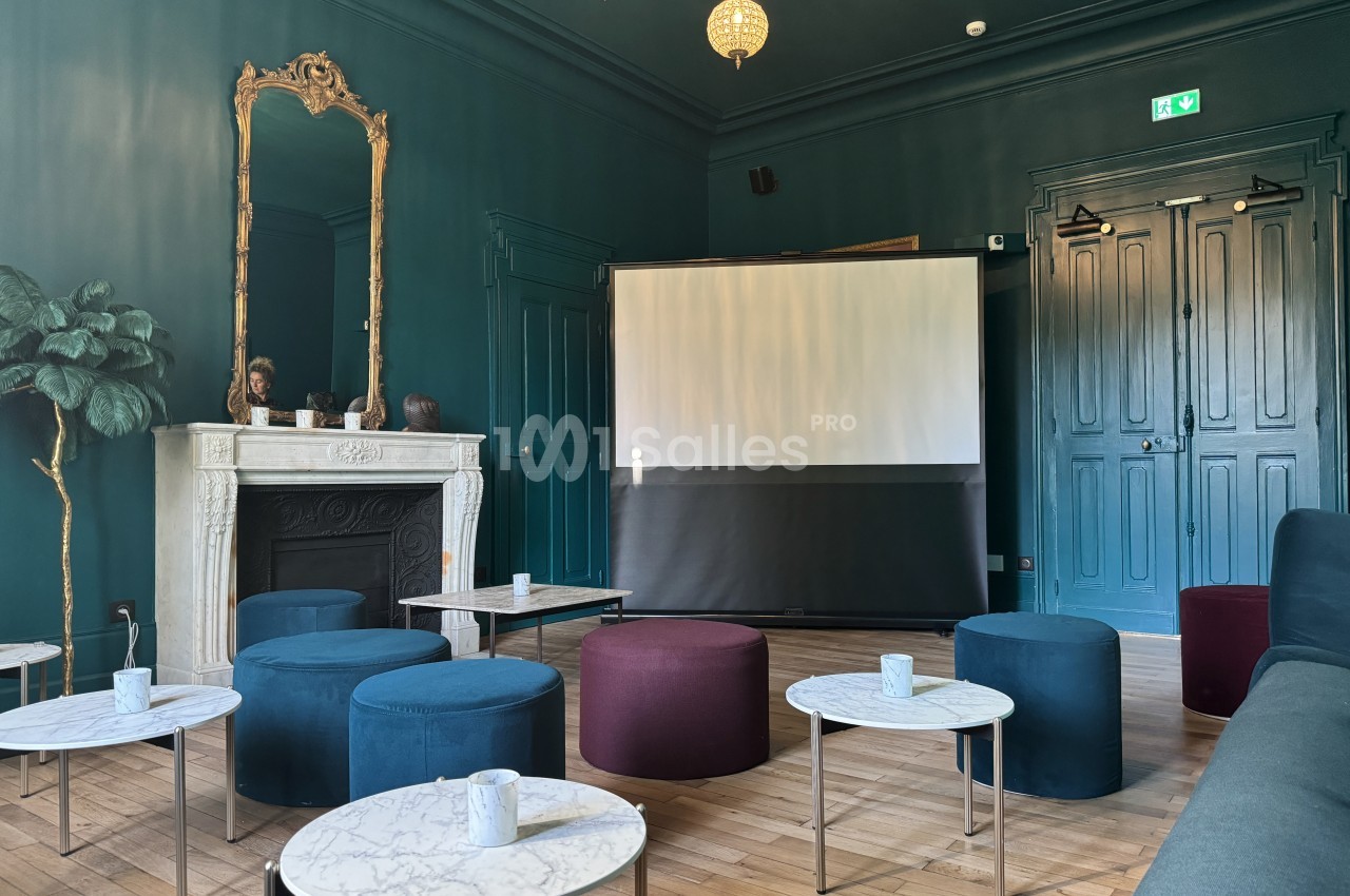 Salle élégante avec murs sombres, cheminée blanche, tables en marbre, poufs colorés et écran de projection.