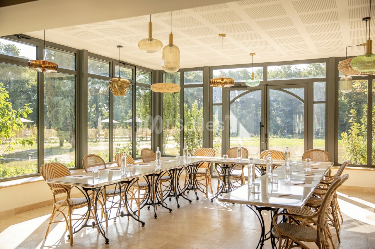 Salle lumineuse avec grandes baies vitrées, tables alignées et chaises en rotin, vue sur un jardin verdoyant.