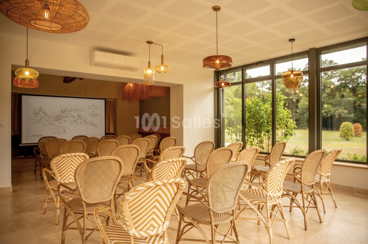 Salle lumineuse avec des chaises en rotin disposées face à un écran de projection, vue sur un jardin à travers de grandes…