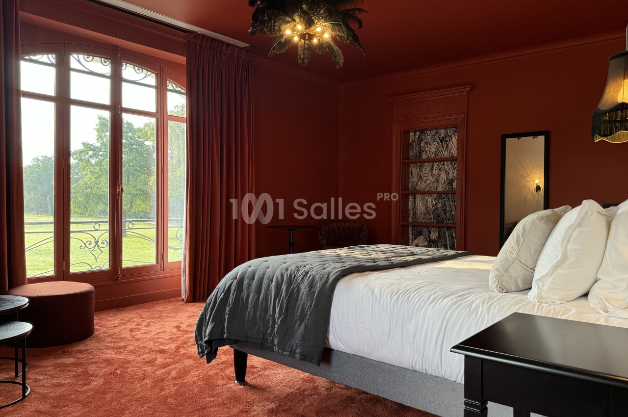 Chambre avec murs rouges, lit double, grande fenêtre donnant sur un jardin, et éclairage décoratif au plafond.