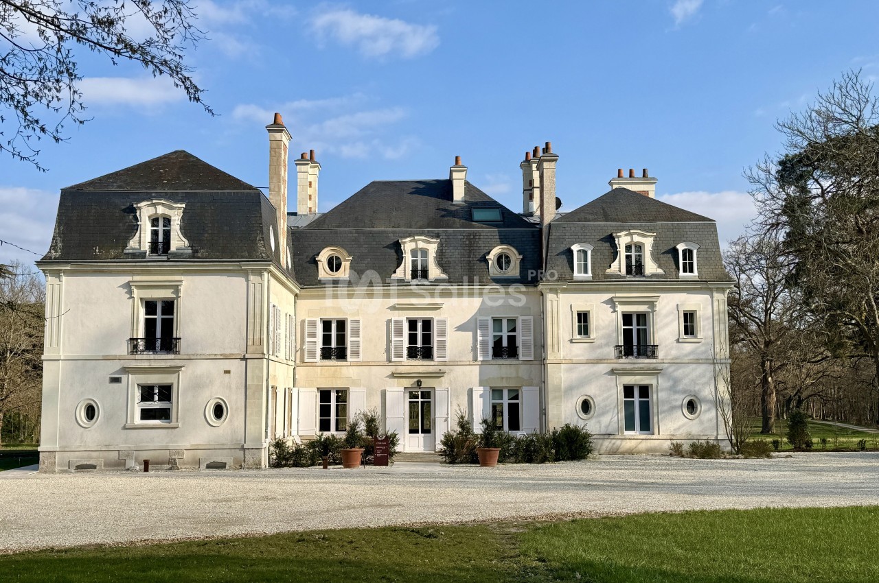 Façade d'un château blanc à deux étages avec toit en ardoise, entouré d'arbres et d'une allée gravillonnée.