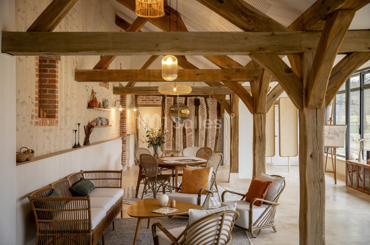 Salle de séjour lumineuse avec poutres en bois apparentes, mobilier en rotin et grandes fenêtres donnant sur l'extérieur.
