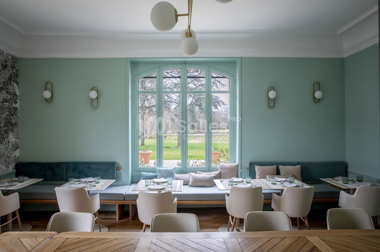 Salle de restaurant lumineuse avec tables dressées, banquettes vertes, chaises blanches et vue sur un jardin arboré.