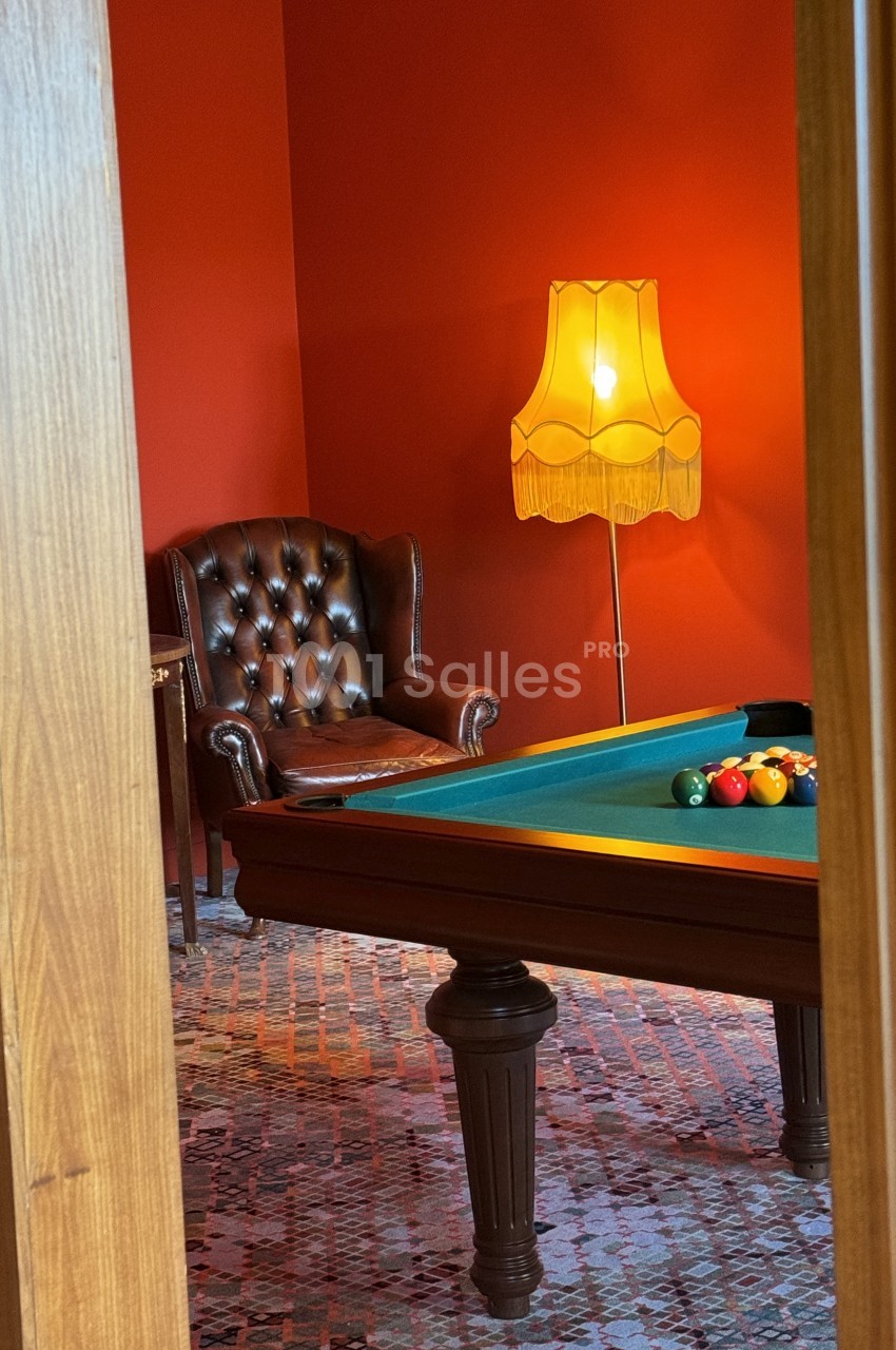 Salle avec un billard, un fauteuil en cuir marron et une lampe rétro éclairant un mur rouge.