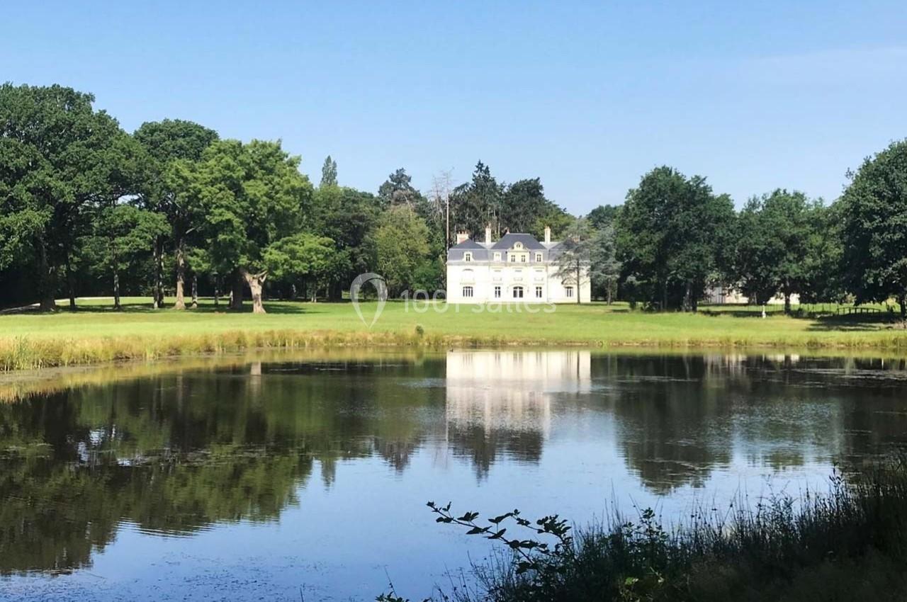 Manoir blanc entouré d'arbres, reflété dans un étang sous un ciel bleu clair.