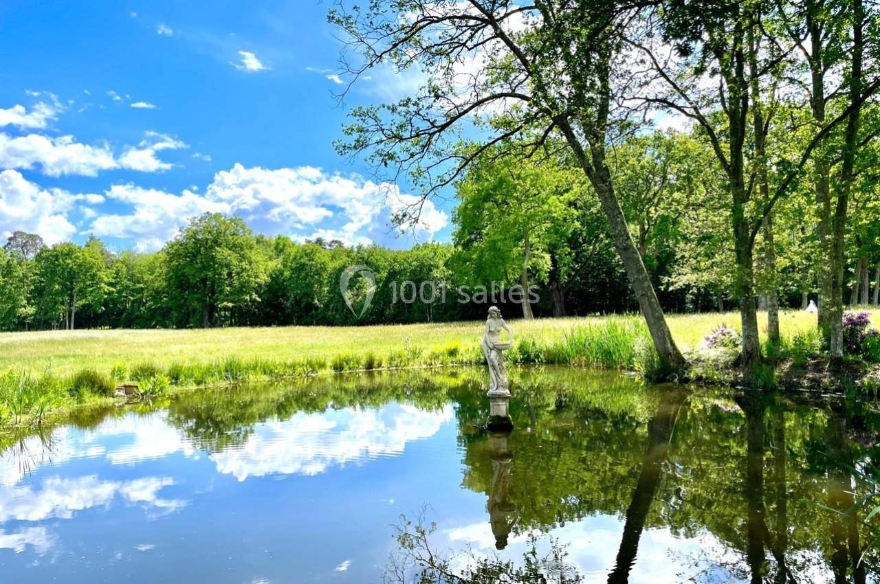 Statue au bord d'un étang entouré d'arbres et d'une prairie sous un ciel bleu avec quelques nuages.