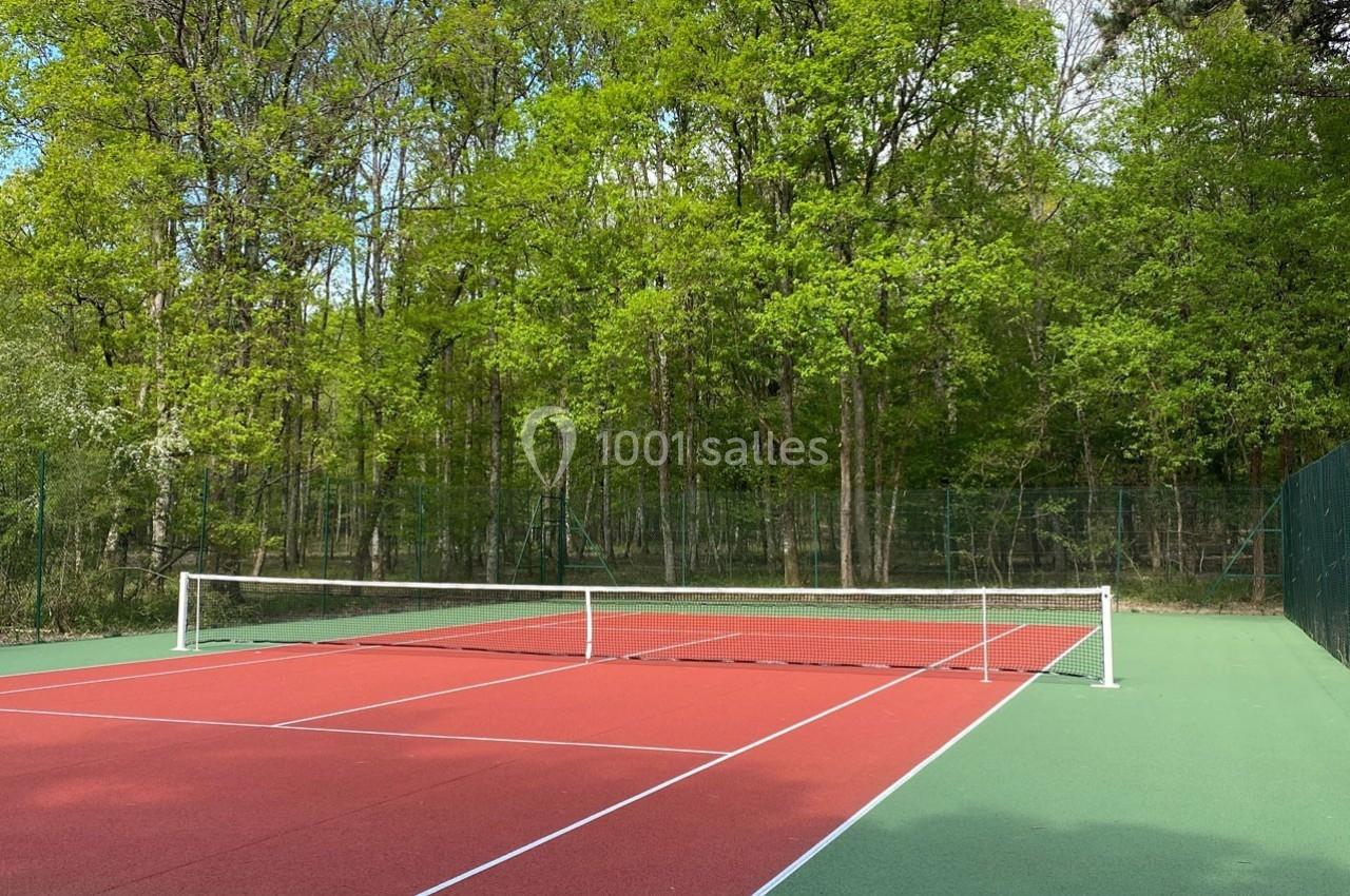 Terrain de tennis extérieur avec revêtement rouge et vert, entouré d'arbres et de végétation dense.