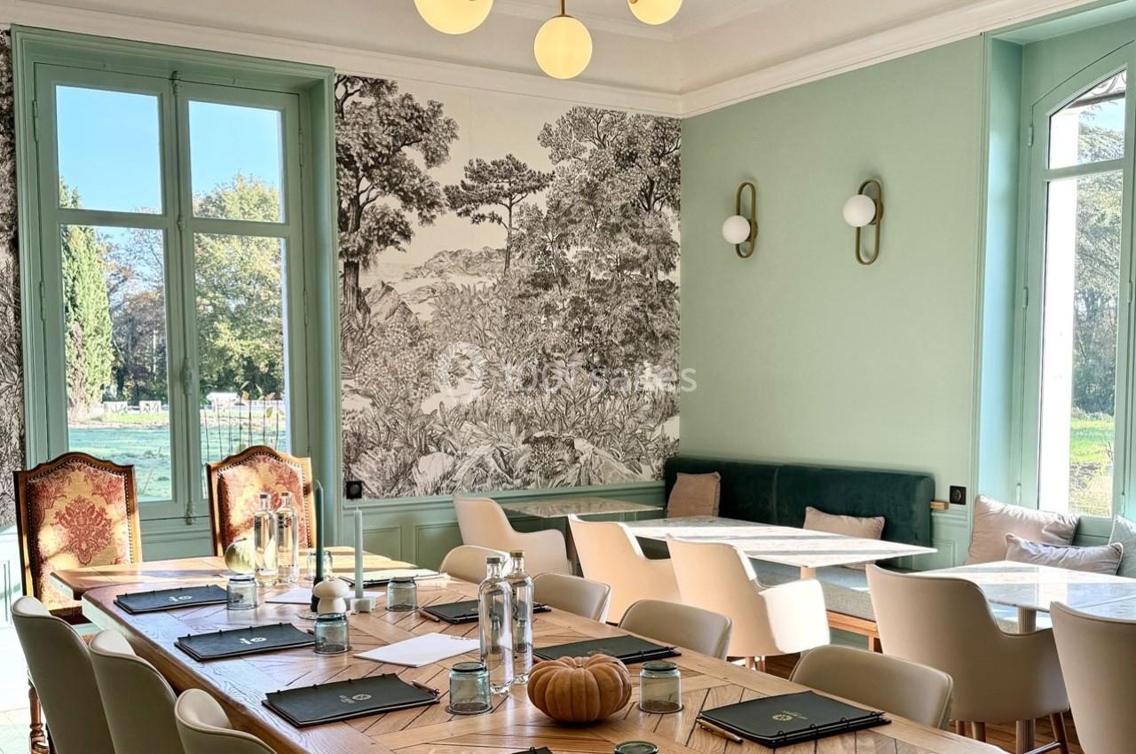 Salle lumineuse avec grande table en bois, chaises modernes, banquette, fresque murale et vue sur un jardin.