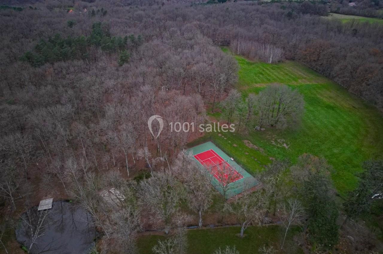Vue aérienne d'un terrain de tennis rouge entouré de forêt, avec une petite mare et des prairies avoisinantes.