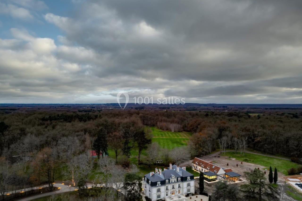 Vue aérienne d'un château entouré de forêts et de prairies sous un ciel nuageux.