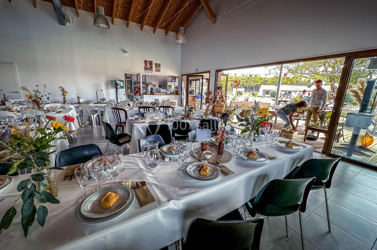 Salle de réception décorée pour un repas, avec tables dressées, fleurs et grandes baies vitrées donnant sur l'extérieur.
