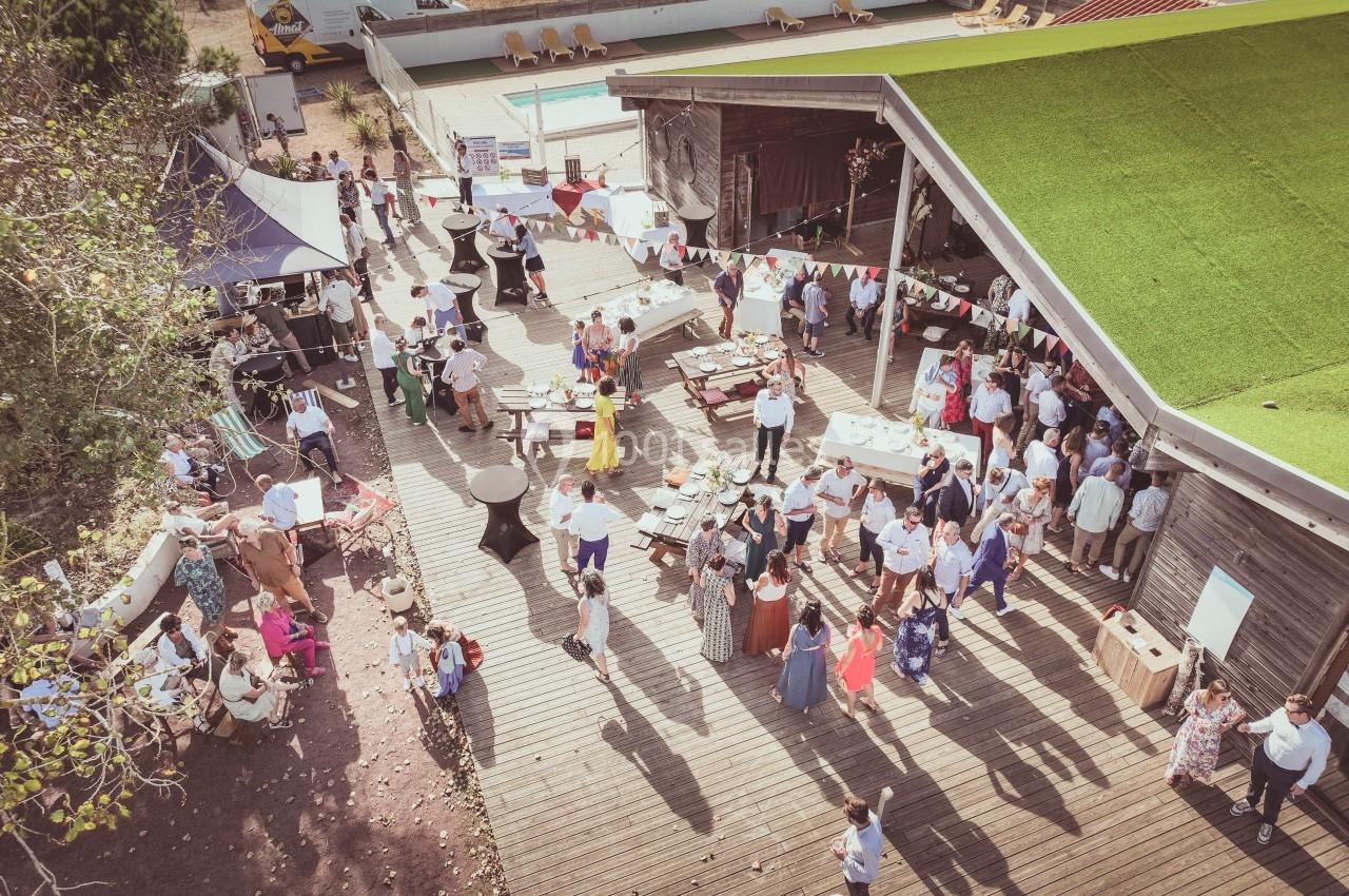 Vue aérienne d'un événement en extérieur avec des invités rassemblés sur une terrasse en bois, décorée de guirlandes.