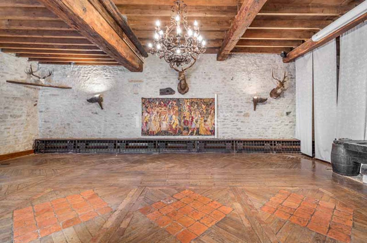 Salle avec murs en pierre, plafond en poutres apparentes, lustre central, tapisseries murales et parquet en bois.