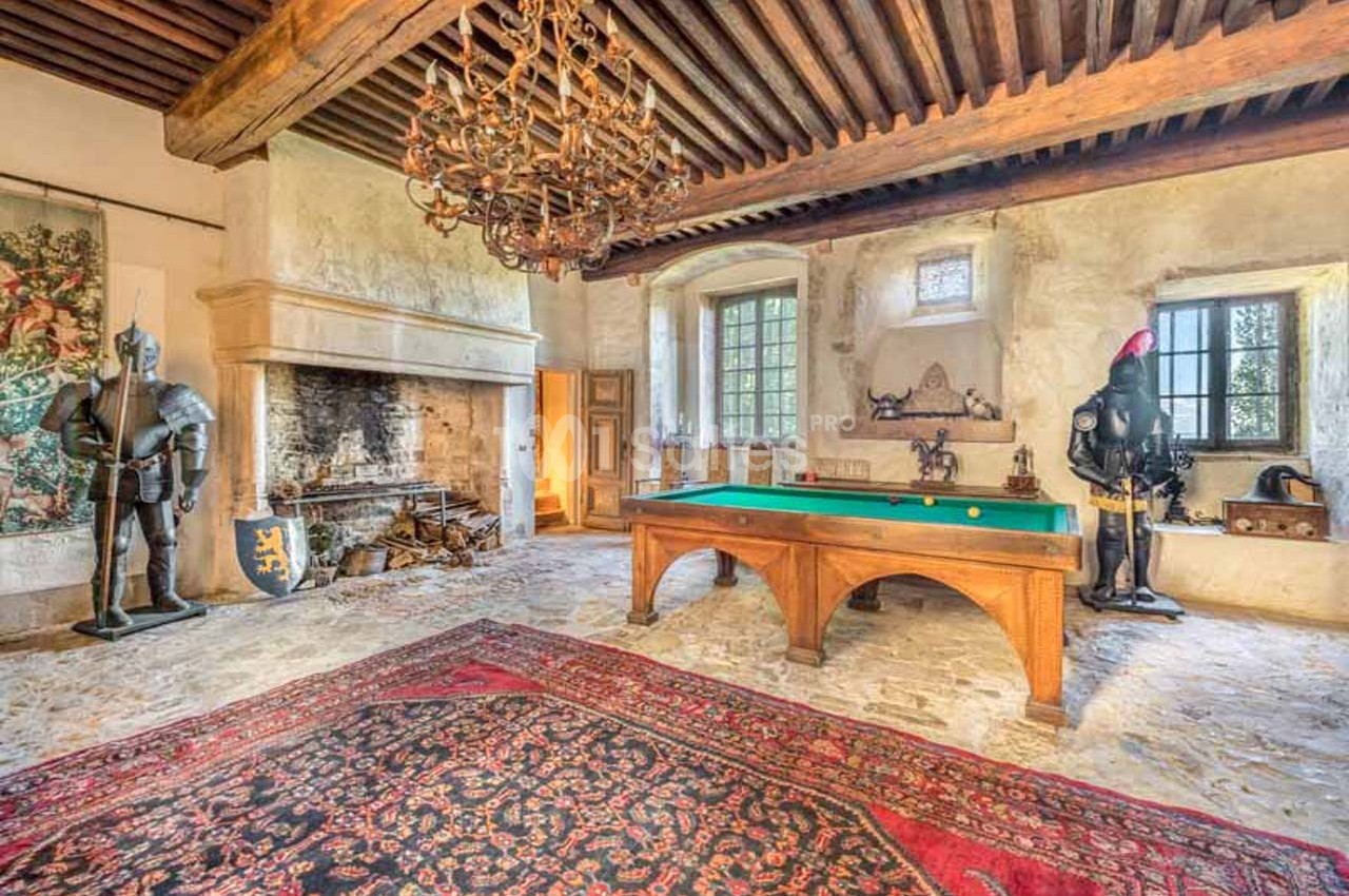 Salle avec billard, cheminée en pierre, tapis ancien, armures médiévales et plafond en poutres apparentes.
