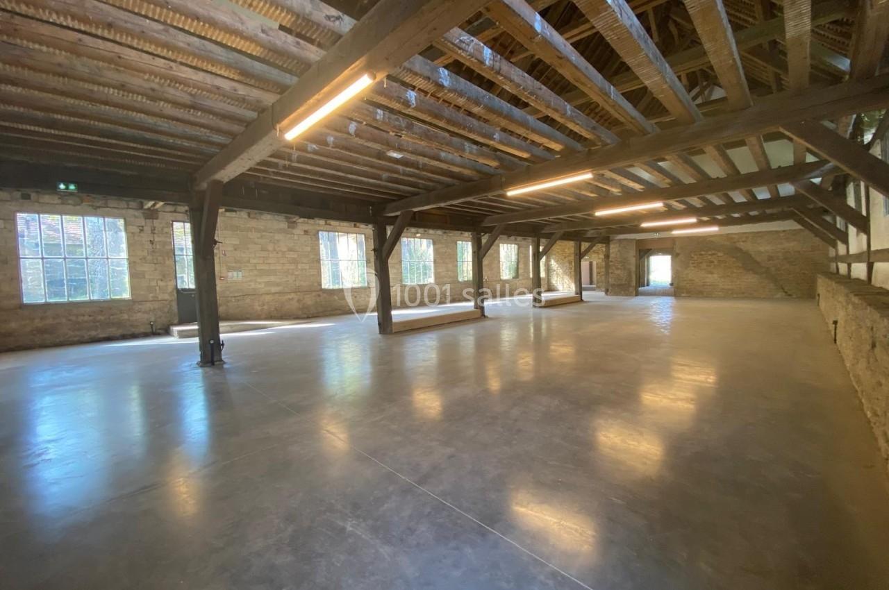 Intérieur d'un grand espace industriel rénové avec poutres en bois, murs en pierre et sol en béton poli.