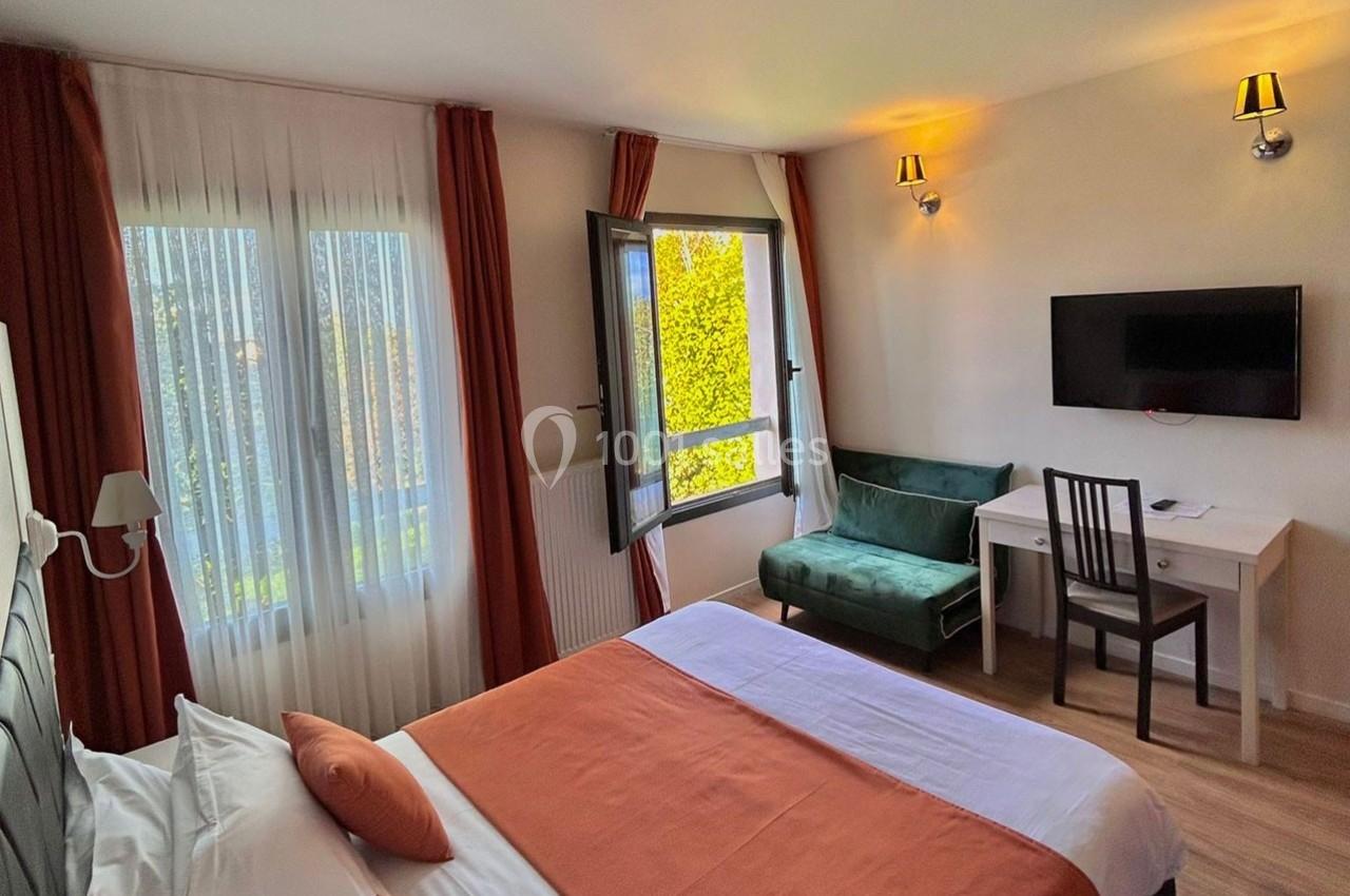 Chambre lumineuse avec lit double, bureau, télévision, canapé vert, et fenêtre ouverte donnant sur un jardin.