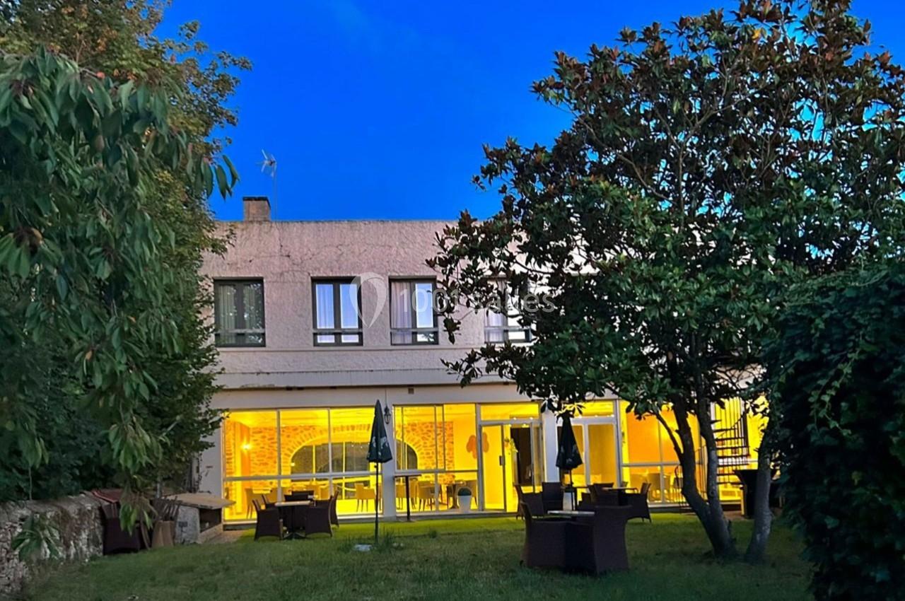 Façade d'un bâtiment avec grandes baies vitrées éclairées, entouré d'arbres et d'un jardin en soirée.
