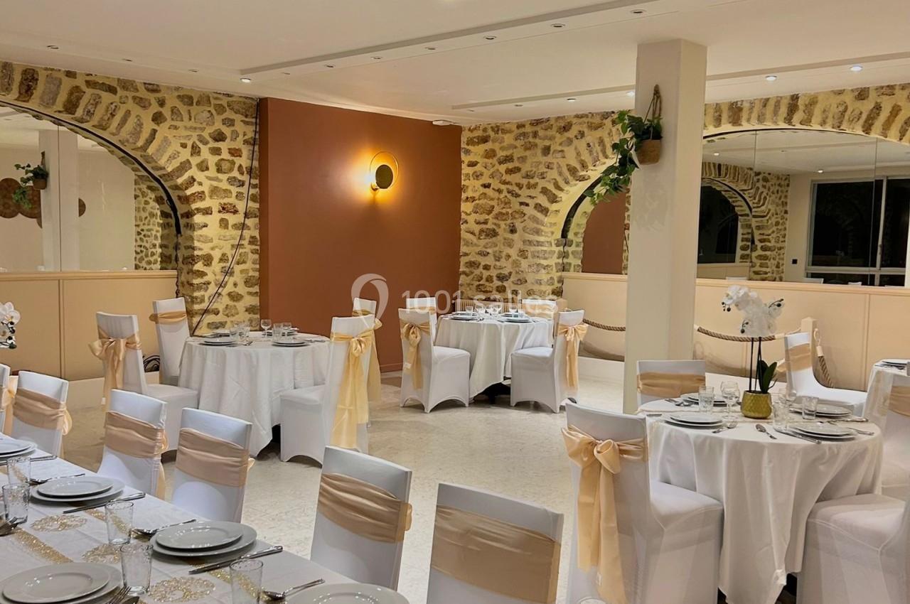 Salle de réception élégante avec tables dressées, nappes blanches, chaises décorées et murs en pierre apparente.