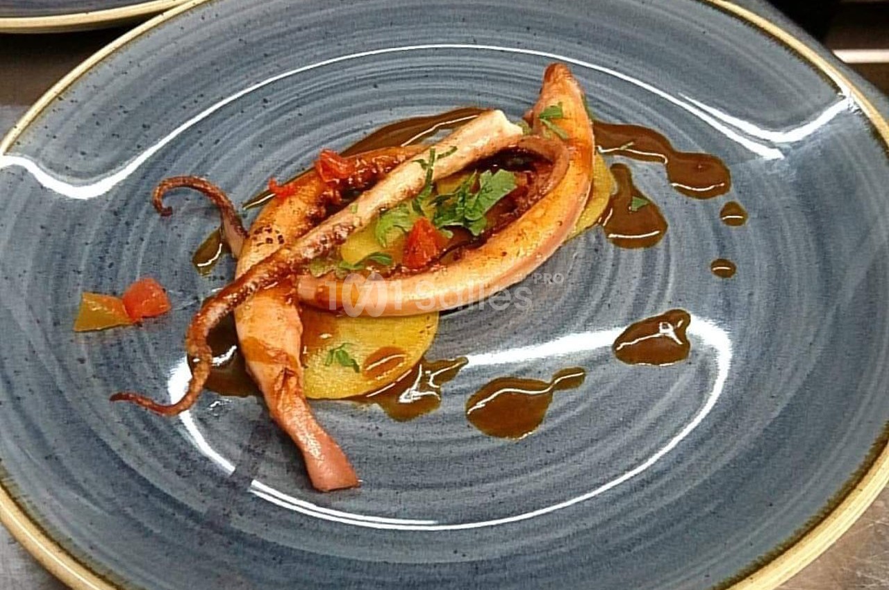 Assiette avec un calamar grillé, accompagné de légumes et d'une sauce disposée en filet sur une assiette bleue.