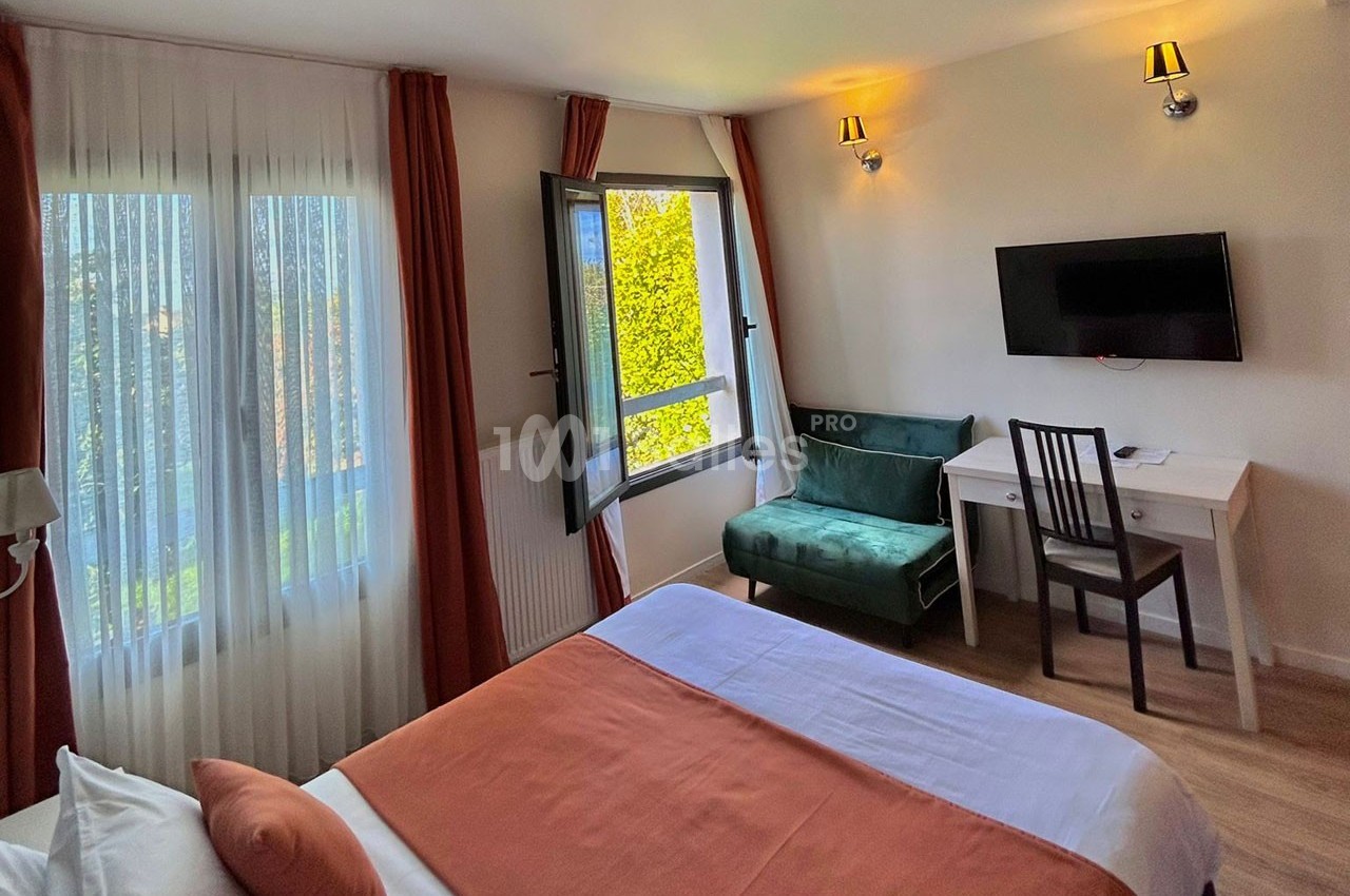 Chambre lumineuse avec lit double, bureau, fauteuil, télévision, fenêtre ouverte et rideaux rouges.