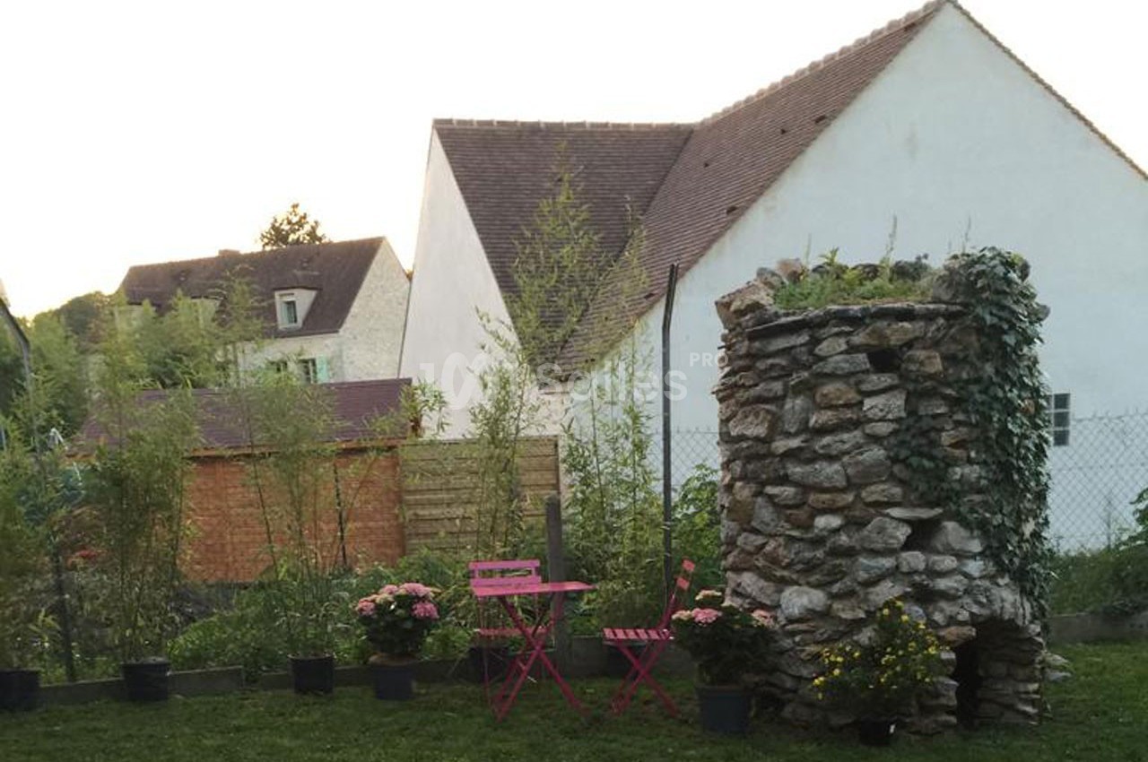 Petit jardin avec une table et des chaises roses, entouré de plantes et d'un puits en pierre devant des maisons.