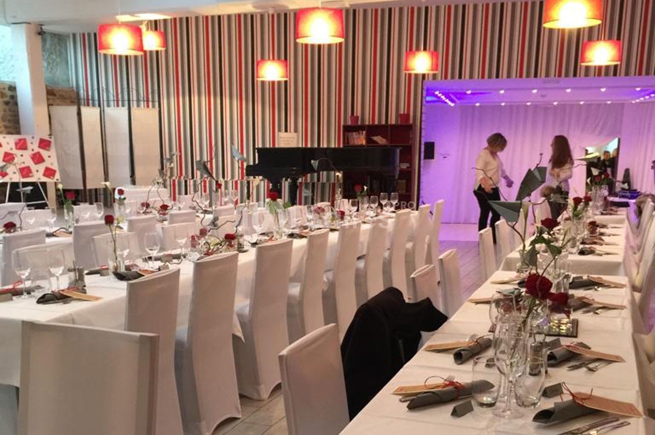 Salle décorée pour un événement avec des tables dressées, des chaises blanches et des lumières suspendues.