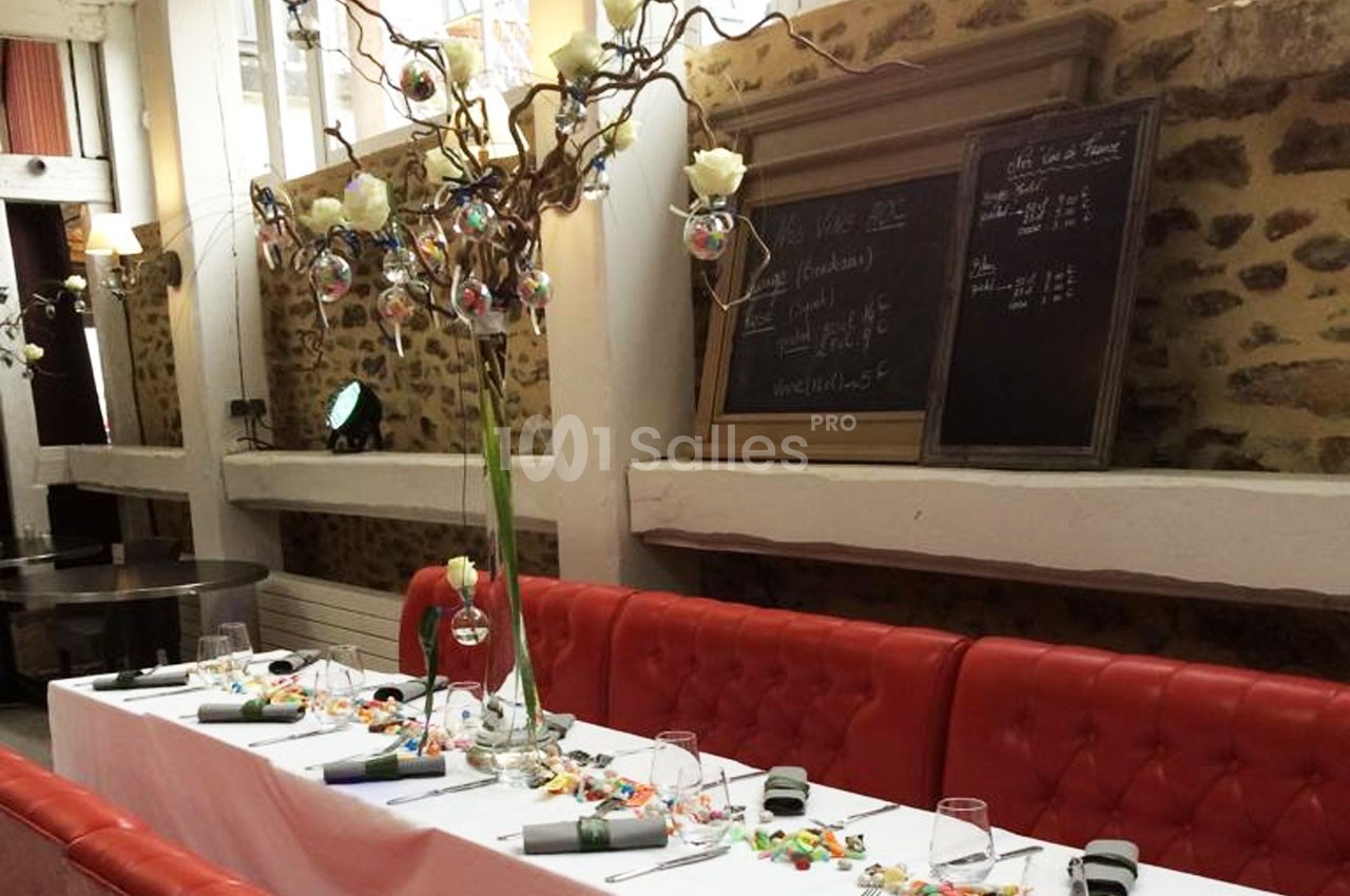 Table dressée avec nappes blanches, décorations florales et bonbons, dans une salle avec murs en pierre et tableau noir.