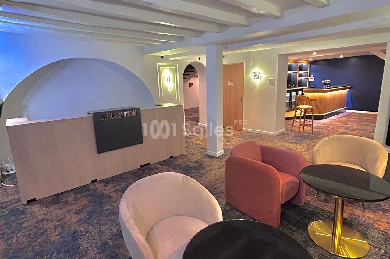 Espace d'accueil avec comptoir de réception, coin salon avec fauteuils et tables, et bar en arrière-plan.