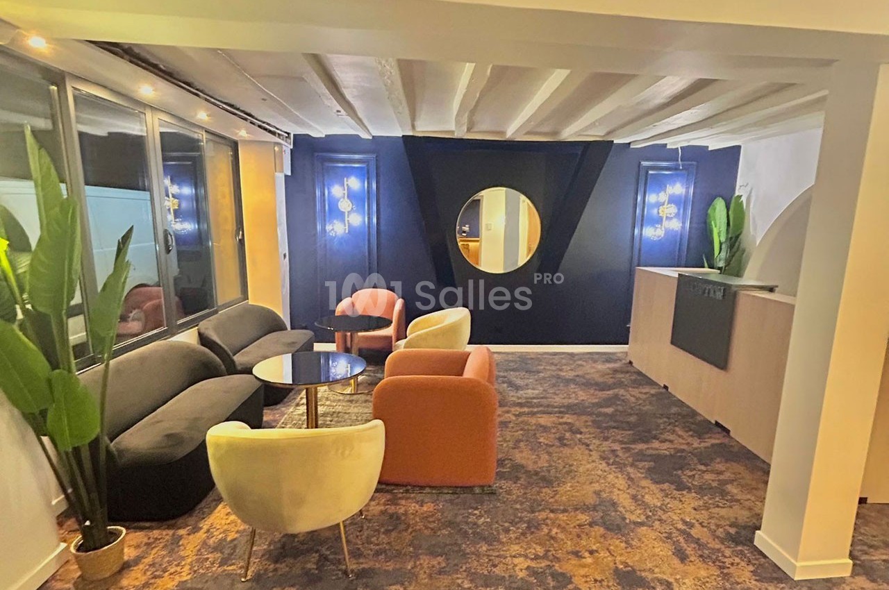 Espace lounge moderne avec fauteuils colorés, table basse, miroir rond et plantes, éclairé par des suspensions.
