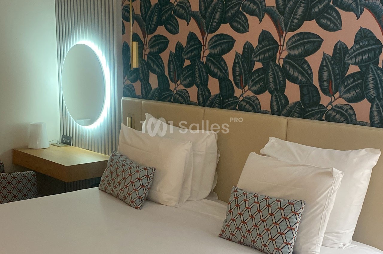 Lit double avec coussins décoratifs, table de chevet, miroir rond lumineux et mur tapissé de motifs végétaux.